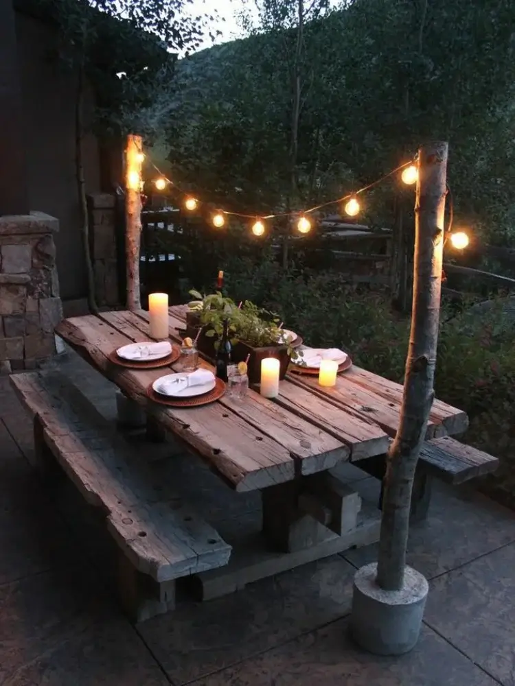 A rustic raw wood picnic table under string lights, set simply with pillar candles and basic plates for a cozy winter dinner.