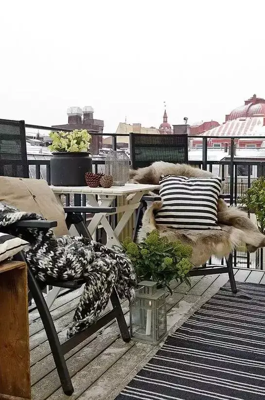 A small winter balcony bistro table set for two with a lantern, pinecones, and chairs draped with soft blankets and cushions.