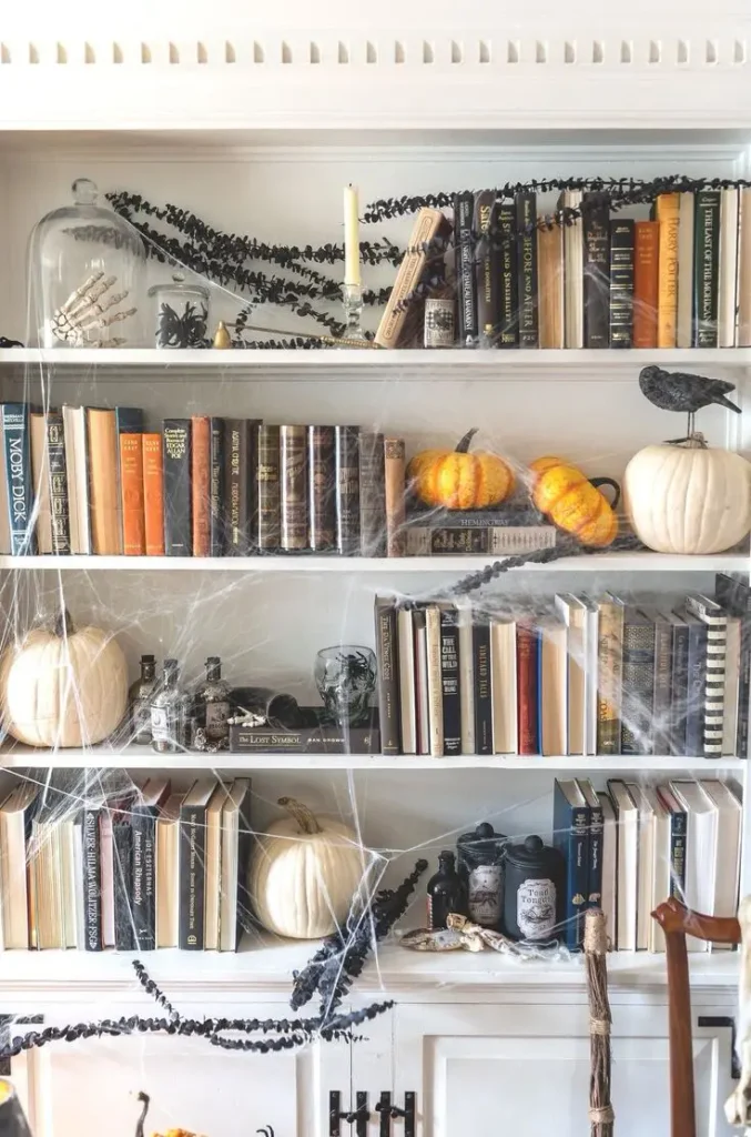 Small apartment Halloween decor on bookshelf with cobwebs, ravens and pumpkins.